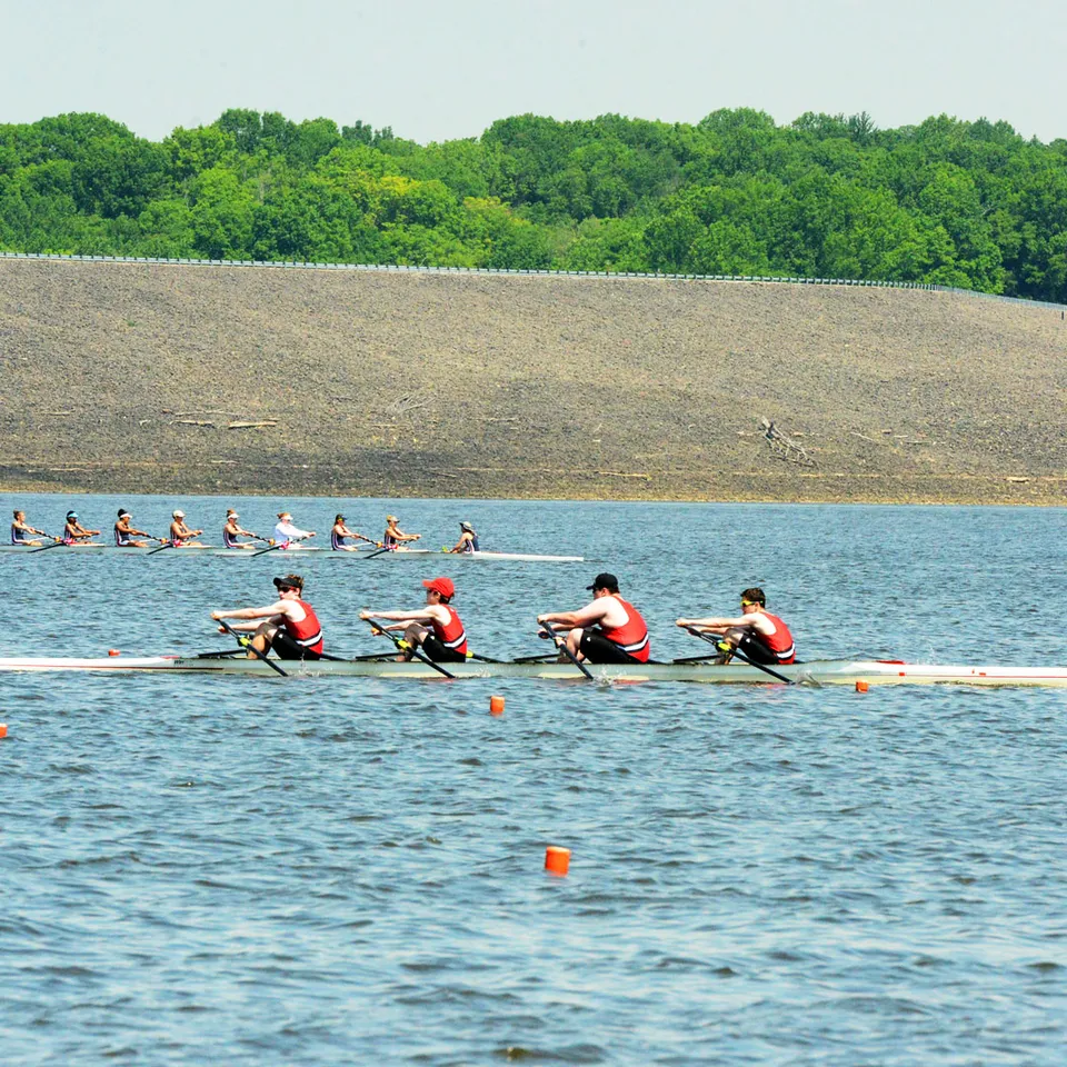 La squadra di canottaggio della Rivermont Collegiate si allena su uno specchio d'acqua con una sponda erbosa e alberi sullo sfondo.