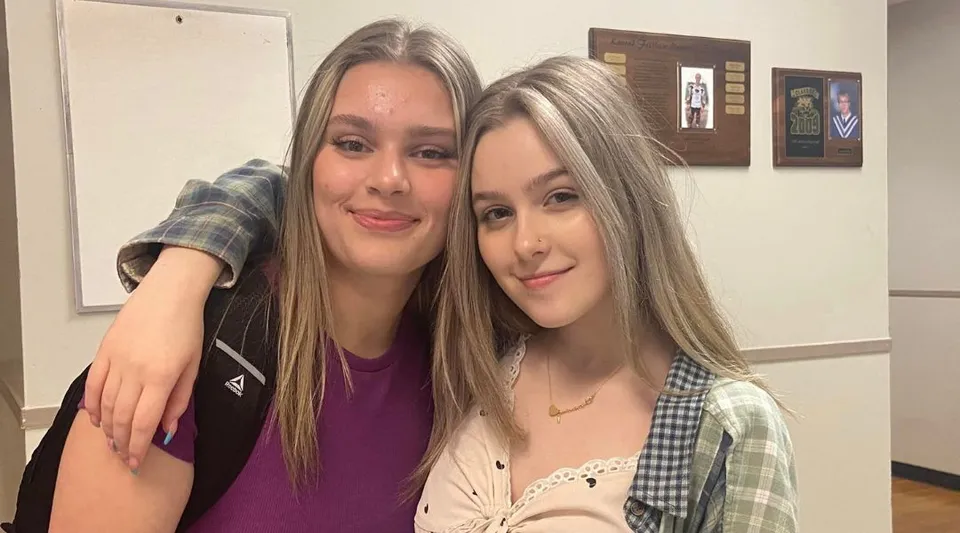 Due studentesse con lunghi capelli biondi posano sorridenti in una stanza con opere d'arte incorniciate nel campus della Lewisporte Collegiate.