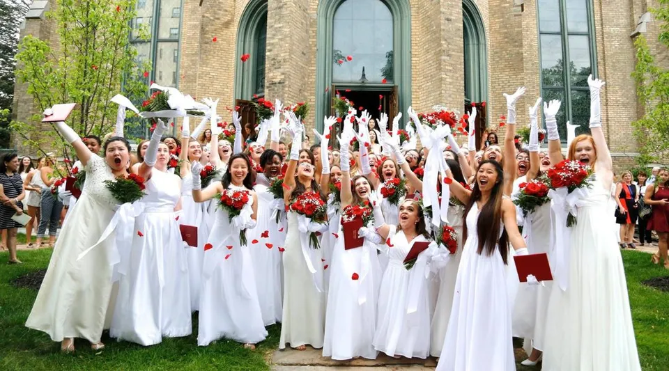 Studenti e studentesse della Buffalo Seminary si radunano in tuniche bianche con fiori rossi davanti a un edificio decorato.