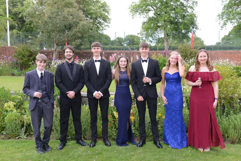 Un gruppo di studenti della Lucton School in abiti formali si trova insieme in un giardino con alberi e aree verdi.