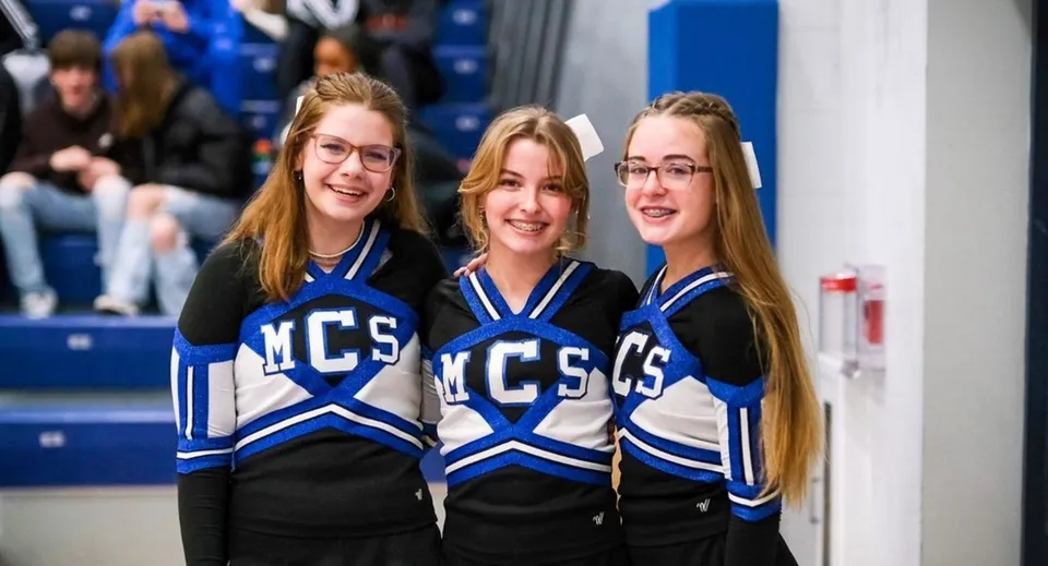 Tre studentesse in divisa da cheerleading con il logo MCS posano davanti a uno sfondo bianco e blu nel campus della Middletown Christian School.