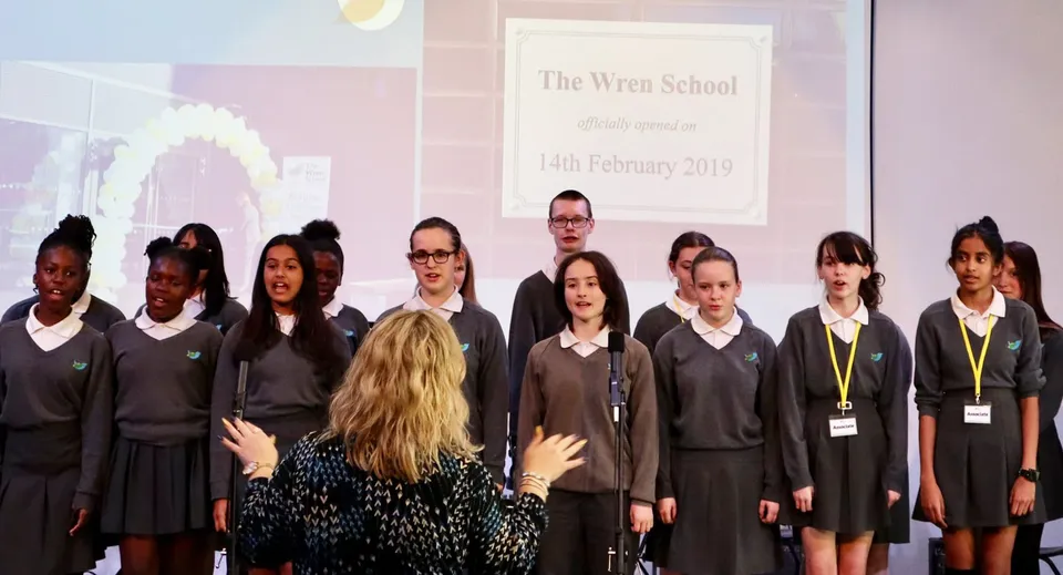 Un coro di studenti della The Wren School canta su un palco davanti a una direttrice e a uno schermo.
