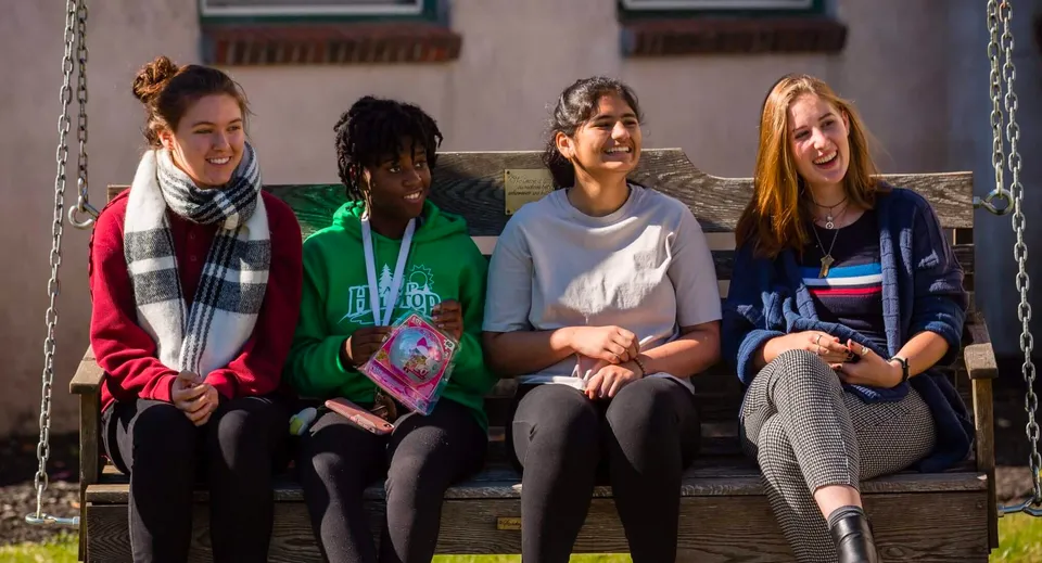 Quattro studentesse siedono su una panchina di legno e chiacchierano nel campus della Stoneleigh-Burnham School.