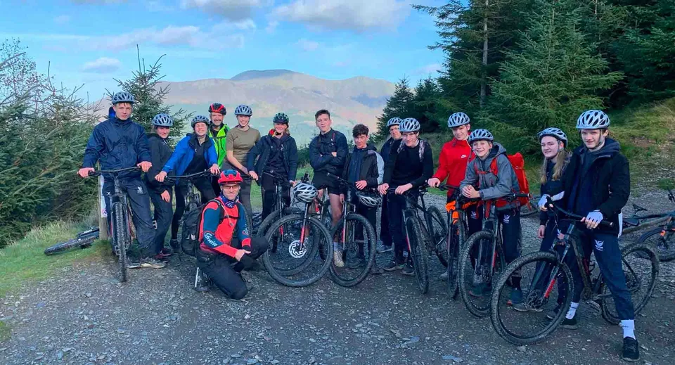Un gruppo di studenti della Stamford Endowed Schools in abbigliamento da ciclista si trova su un sentiero nel bosco con pini e montagne sullo sfondo.