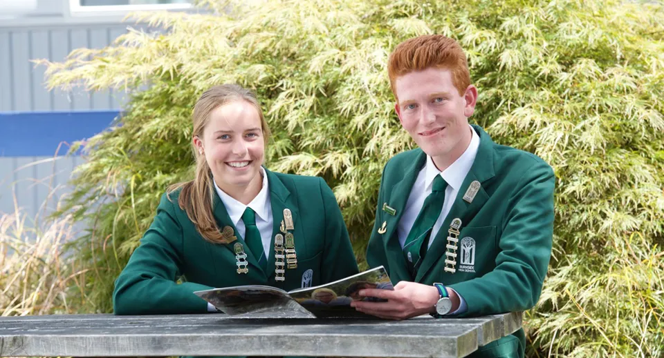 Due studenti in uniforme scolastica verde sono insieme nel campus della Burnside High School davanti a un fogliame verde.