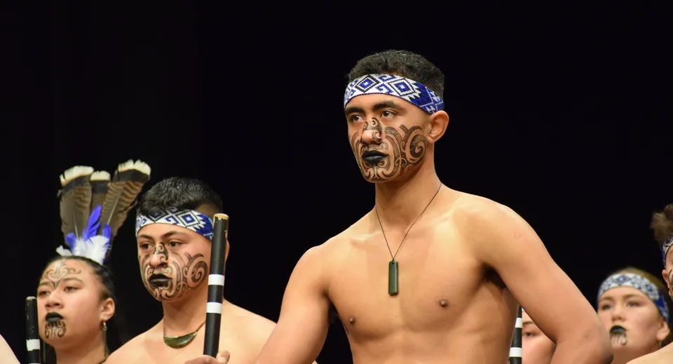 Un gruppo di uomini con tradizionali tatuaggi facciali Maori e copricapi presenta una performance di danza sul palco della King's High School.
