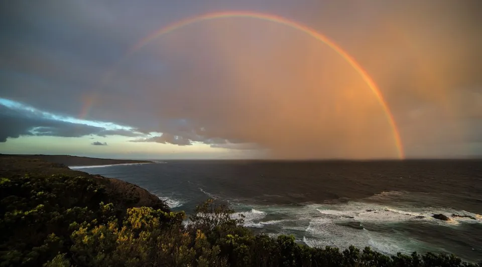 Un arcobaleno si estende sul paesaggio costiero adiacente alla Kingscliff High School, tra rocce scoscese e vegetazione.