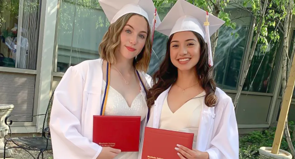 Due studentesse della Woodlands Academy of the Sacred Heart in abito da cerimonia tengono con orgoglio i loro diplomi davanti a un edificio con area verde.