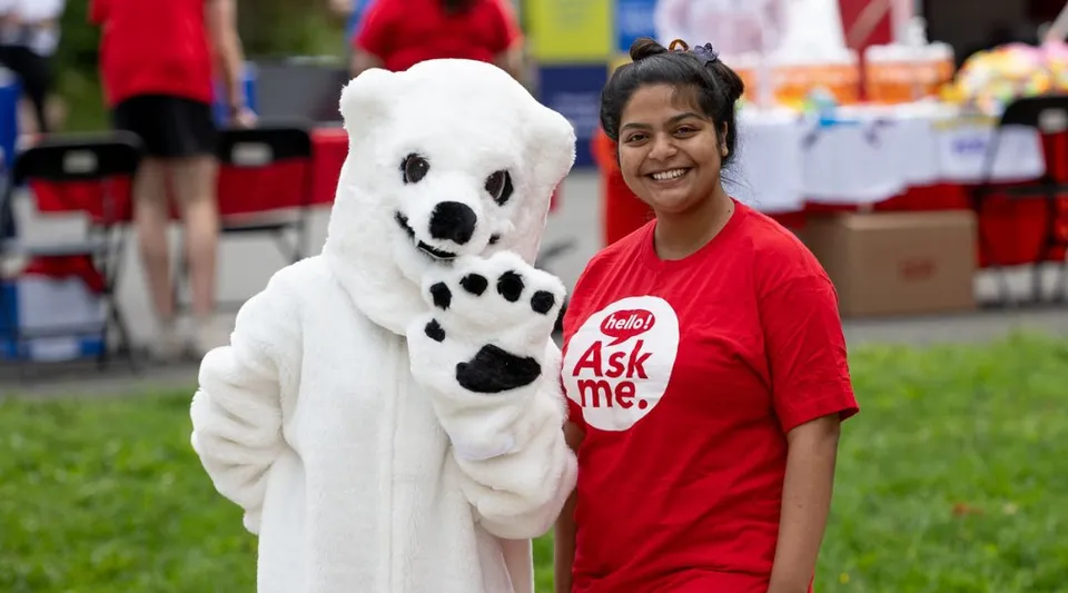 Una persona sorridente con una t-shirt rossa con scritto Ask me si trova accanto a un grande peluche bianco con impronte di zampe nere all'interno del campus della Memorial University.