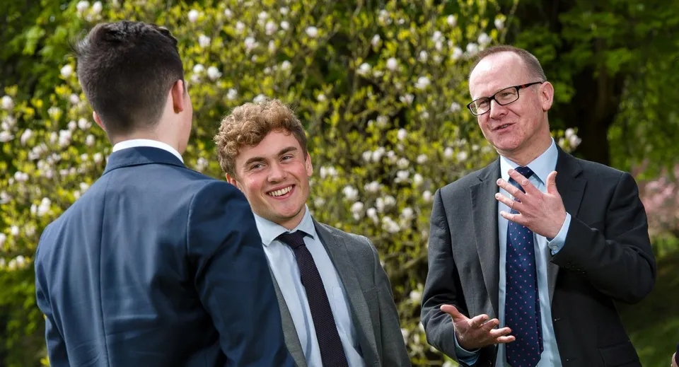 Tre uomini in abito conversano davanti a uno sfondo verde con alberi nel terreno della Kingswood School.