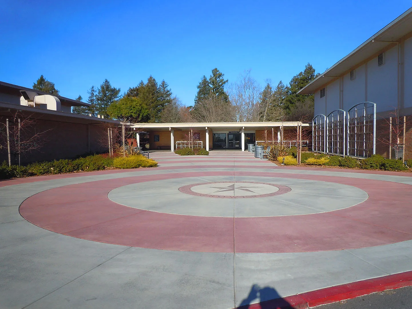 El patio adoquinado con un patrón circular en el campus de Justin Siena High School está rodeado de edificios con grandes ventanas y pinos.