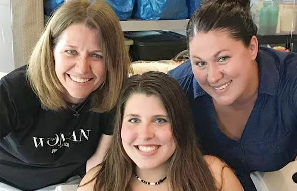 Una joven sonríe rodeada por dos mujeres que se inclinan hacia ella para la foto