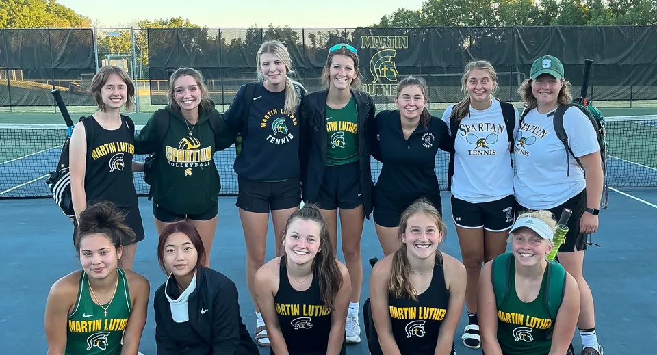 Un gruppo di studentesse della Martin Luther High School posa insieme sul campo da tennis davanti a una recinzione con alberi sullo sfondo.