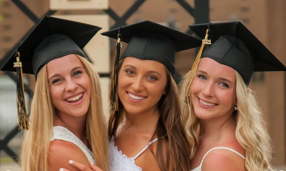 Tre studentesse della Mt. Vernon High School in abiti da diploma posano davanti a uno sfondo in legno.