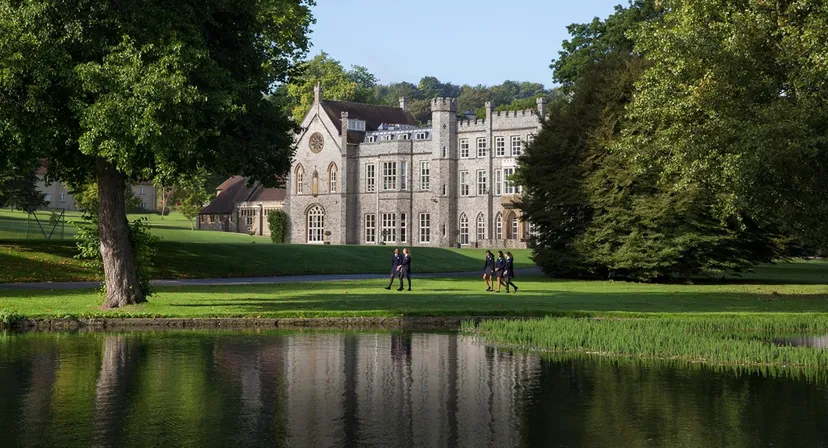 L'edificio storico della Wycombe Abbey School circondato da un parco con un laghetto e studenti sul prato.