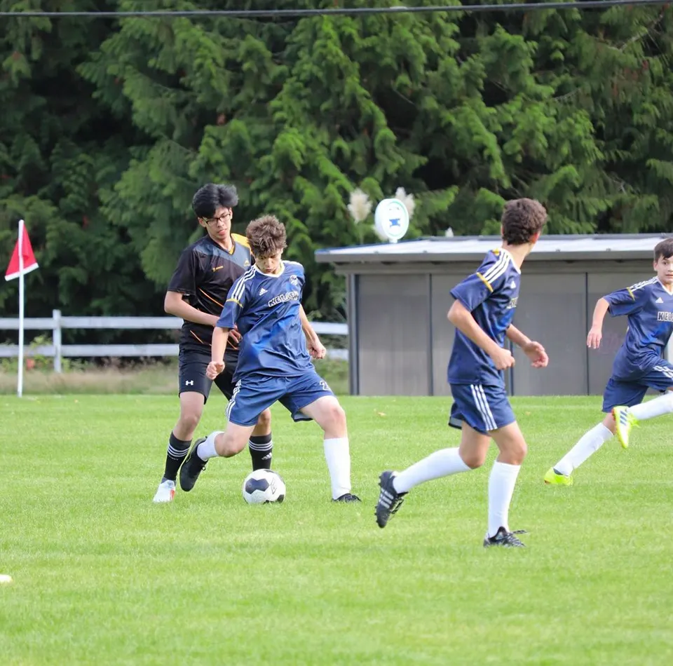 Studenti della Frances Kelsey Secondary School giocano a calcio su un campo erboso con un edificio e alberi sullo sfondo.