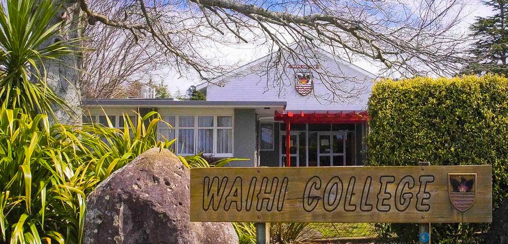 Un grande cartello con la scritta Waihi College è circondato dal verde davanti a un edificio con la tettoia rossa.