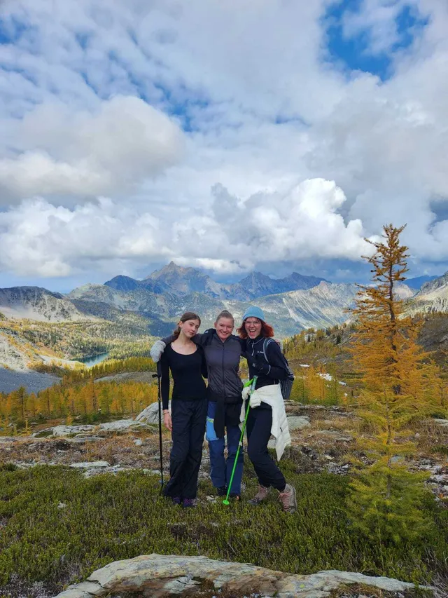 Tre ragazze posano su una cima con larici dorati e le montagne sullo sfondo