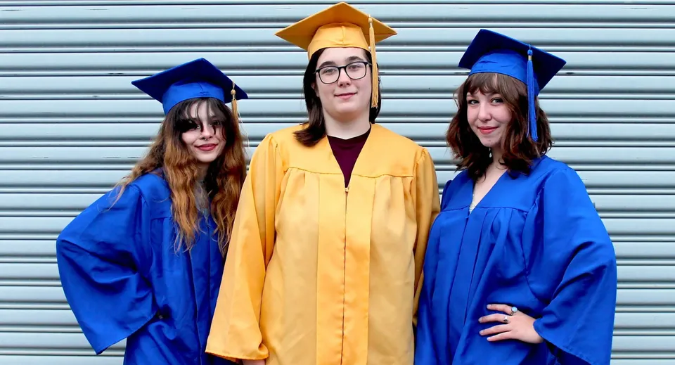 Tre persone in diverse toghe da diploma sono in piedi davanti a una serranda metallica nel campus del Castlepollard Community College.