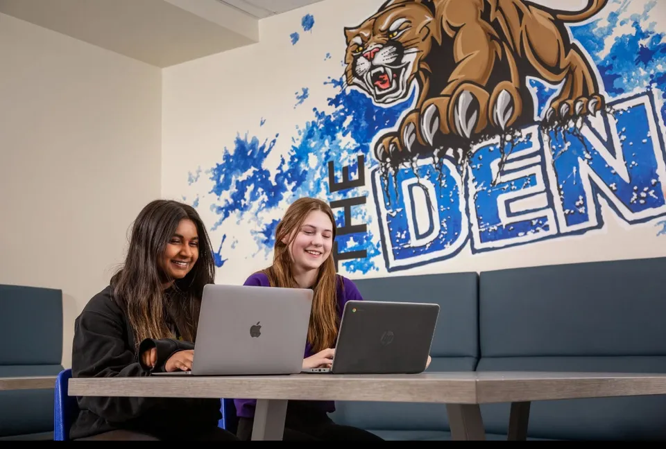 Due studentesse della Covenant Christian School siedono a un tavolo e lavorano al laptop davanti a un murale raffigurante una tigre.