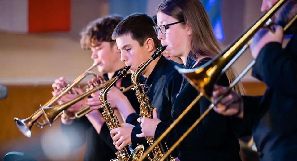 Un gruppo di musicisti suona diversi strumenti a fiato e ottoni sul palco della Felsted School su uno sfondo colorato.