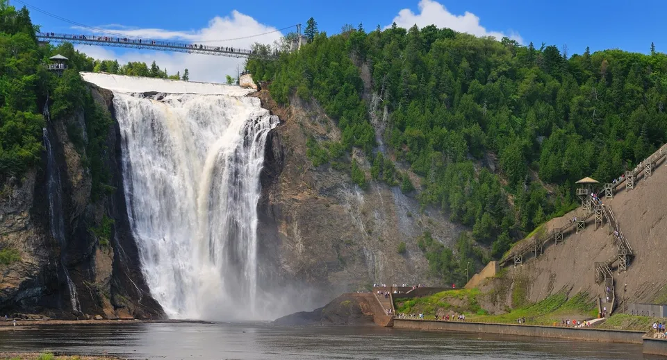 Una grande cascata scende lungo una parete rocciosa con un ponte sospeso in un paesaggio boscoso.