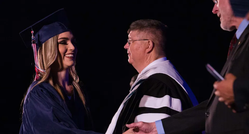 Una giovane diplomata della Millfield School stringe la mano a un uomo anziano in toga accademica durante una cerimonia.