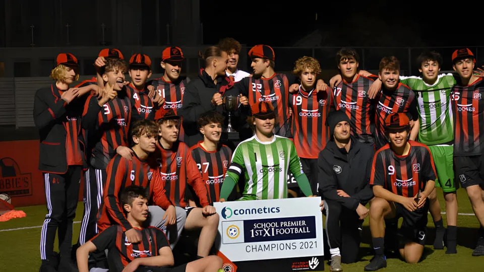 Un gruppo di giovani calciatori con maglie rosse e nere festeggia la vittoria del campionato sul campo da gioco del St. Bede's College.