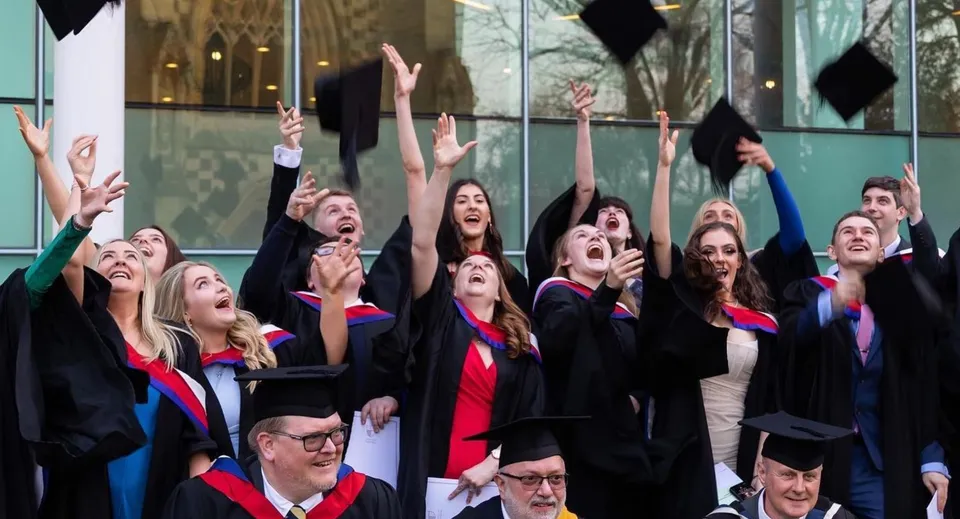 Un gruppo di diplomati della Charterhouse School festeggia il diploma a braccia alzate davanti a un edificio in vetro.