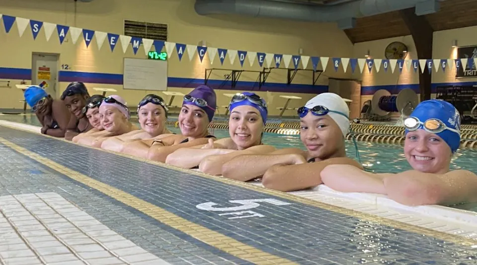 Un gruppo di studentesse sorridenti della Salem Academy siede al bordo di una luminosa piscina al coperto con decorazioni blu e bianche.
