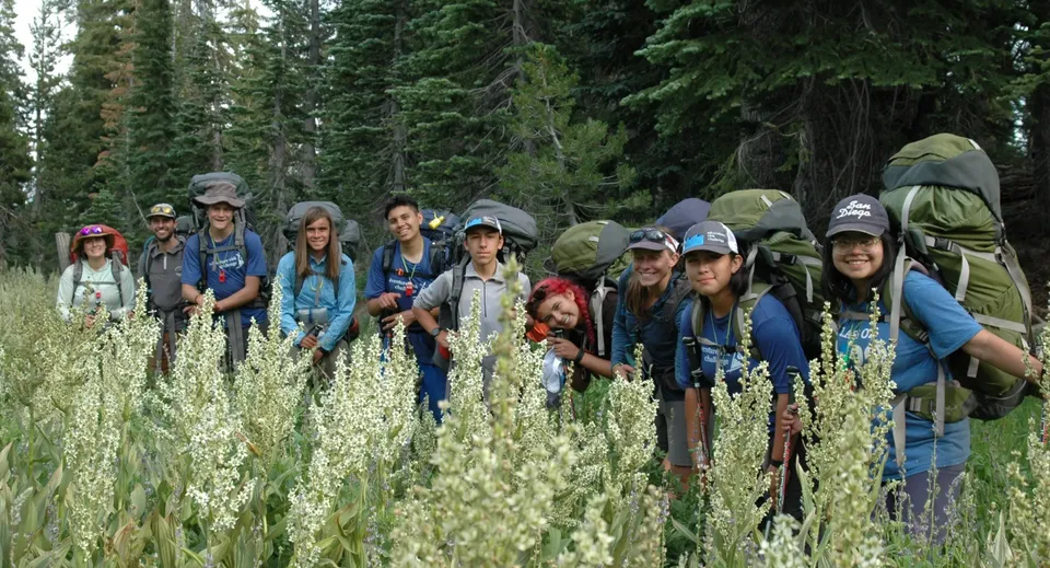 Un gruppo di studenti della Charterhouse School in abbigliamento da trekking si trova su un prato con erba alta e fiori selvatici davanti a un fitto bosco.