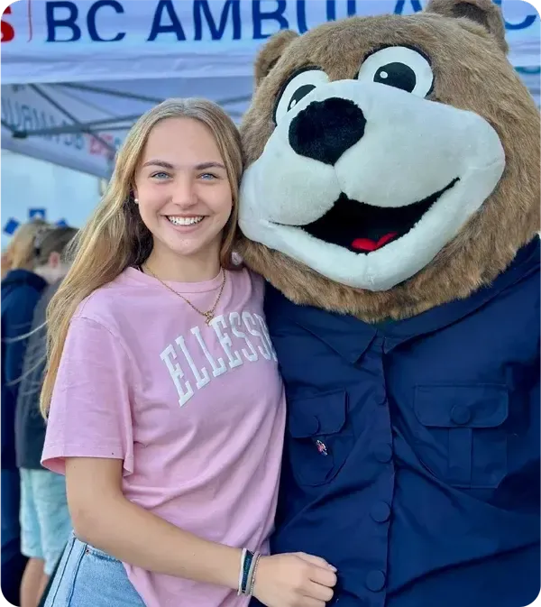 Estudiante posando junto a una mascota disfrazada de oso en una feria