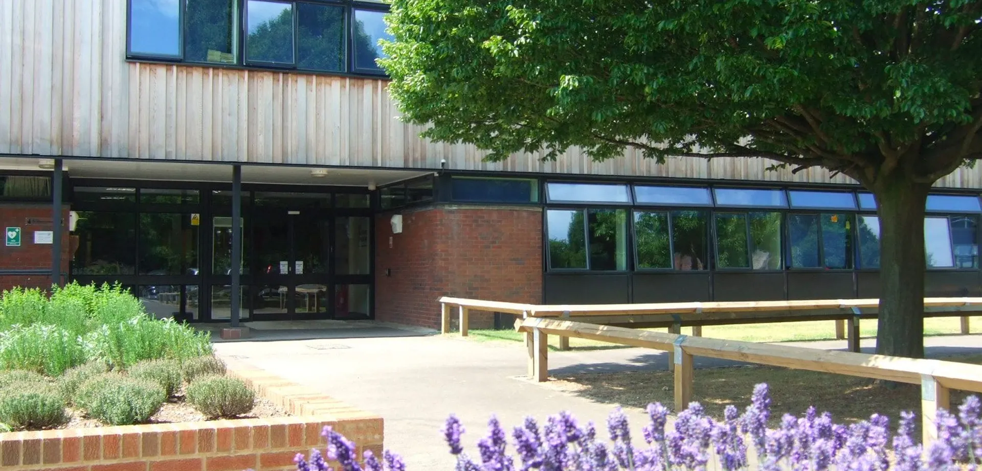 L'edificio scolastico moderno della Denefield School è circondato da piante verdi con fiori lilla e una panchina di legno.