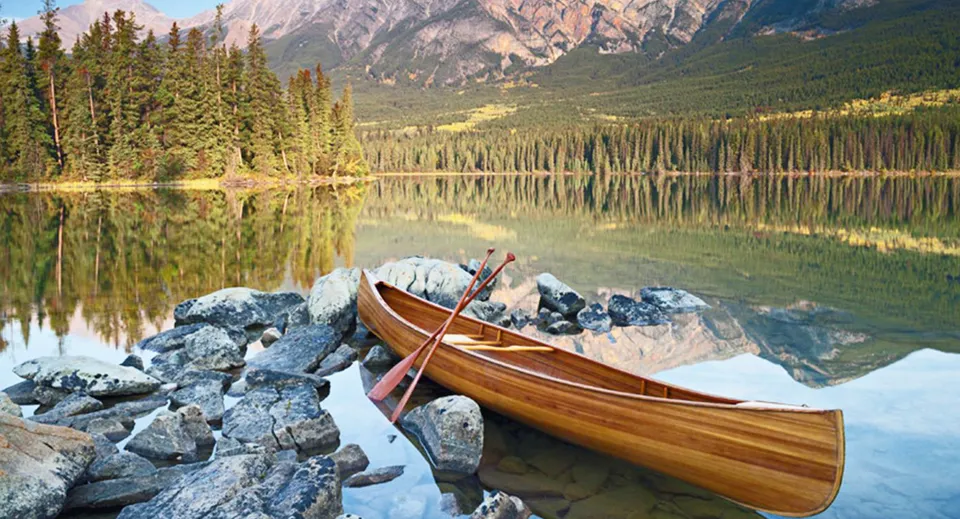 Una canoa in legno si trova sulla riva pietrosa di un lago calmo con montagne e boschi nei dintorni della Eastside Secondary School.