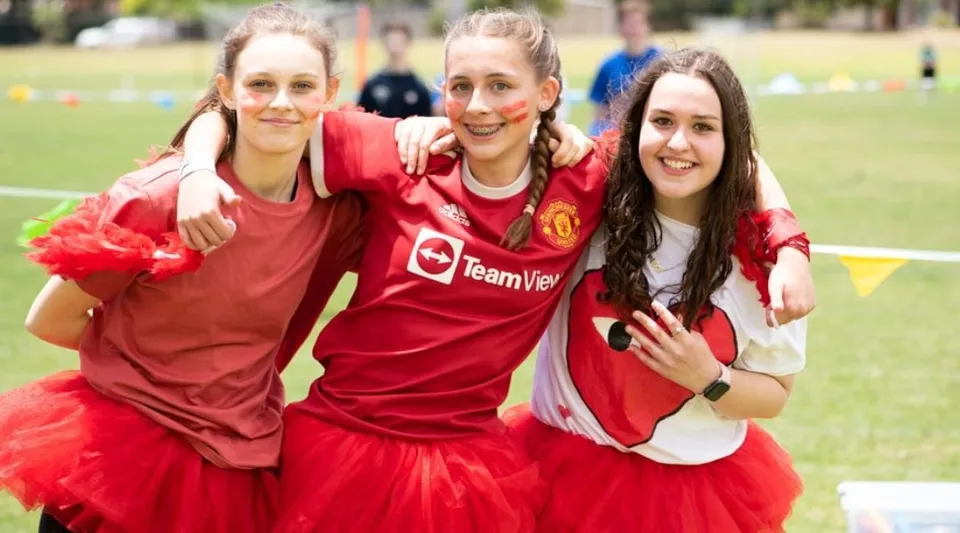 Tre studentesse della Seaview High School indossano maglie rosse del Manchester United e si abbracciano su un campo in erba.