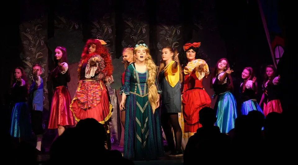 Donne in costumi colorati sul palco della Palmerstown Community School durante un'esibizione teatrale.