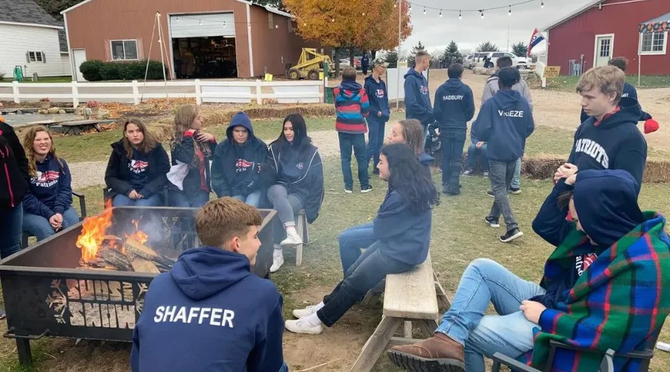 Gli studenti della Cadillac Heritage Christian School si radunano all'aperto in autunno attorno a un braciere con fogliame colorato sullo sfondo.