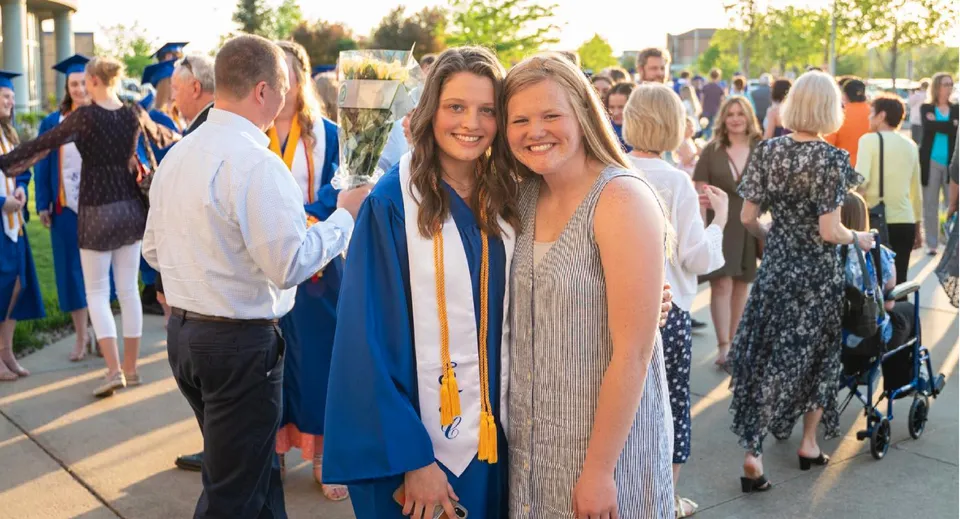 Due studentesse della Sioux Falls Christian School sorridono e posano insieme durante una cerimonia di diploma all'aperto davanti a edifici e spazi verdi.