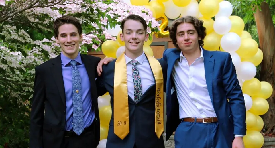 Tre studenti in abito formale sono in piedi davanti ad alberi in fiore e palloncini gialli nel campus della Spectrum Community Secondary School.