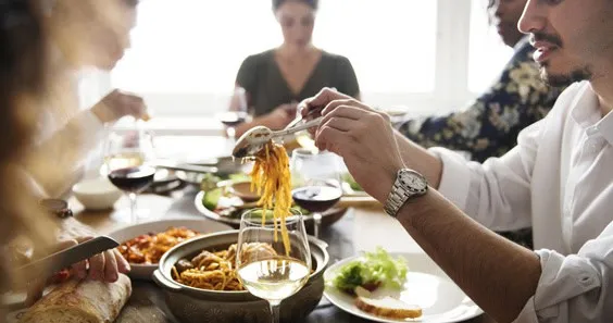 Un hombre sirve pasta con pinzas en una mesa llena de comida y copas de vino