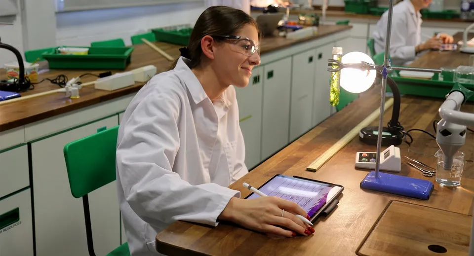 Una persona in camice bianco da laboratorio lavora al tablet in un laboratorio della Queen Anne’s School con strumenti scientifici sullo sfondo.