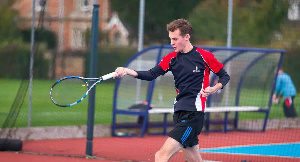Uno studente della Taunton School International gioca a tennis su un campo all'aperto colorato con edifici e alberi sullo sfondo.