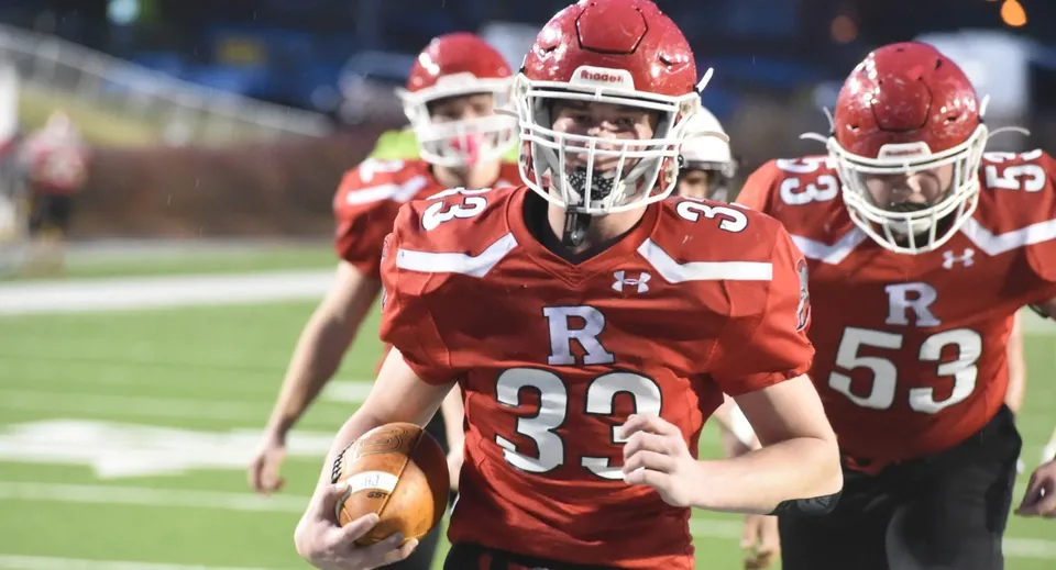 Giocatori di football americano della Walpole High School in maglia rossa corrono con la palla sul campo.