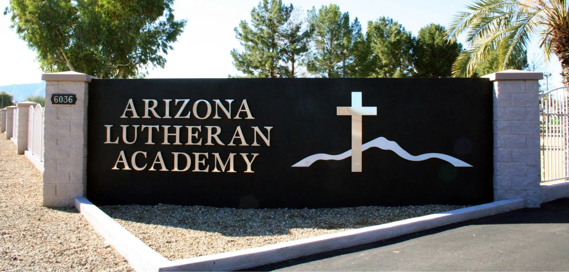 Un grande cartello nero con la scritta Arizona Lutheran Academy e una croce sta davanti a delle palme verdi.