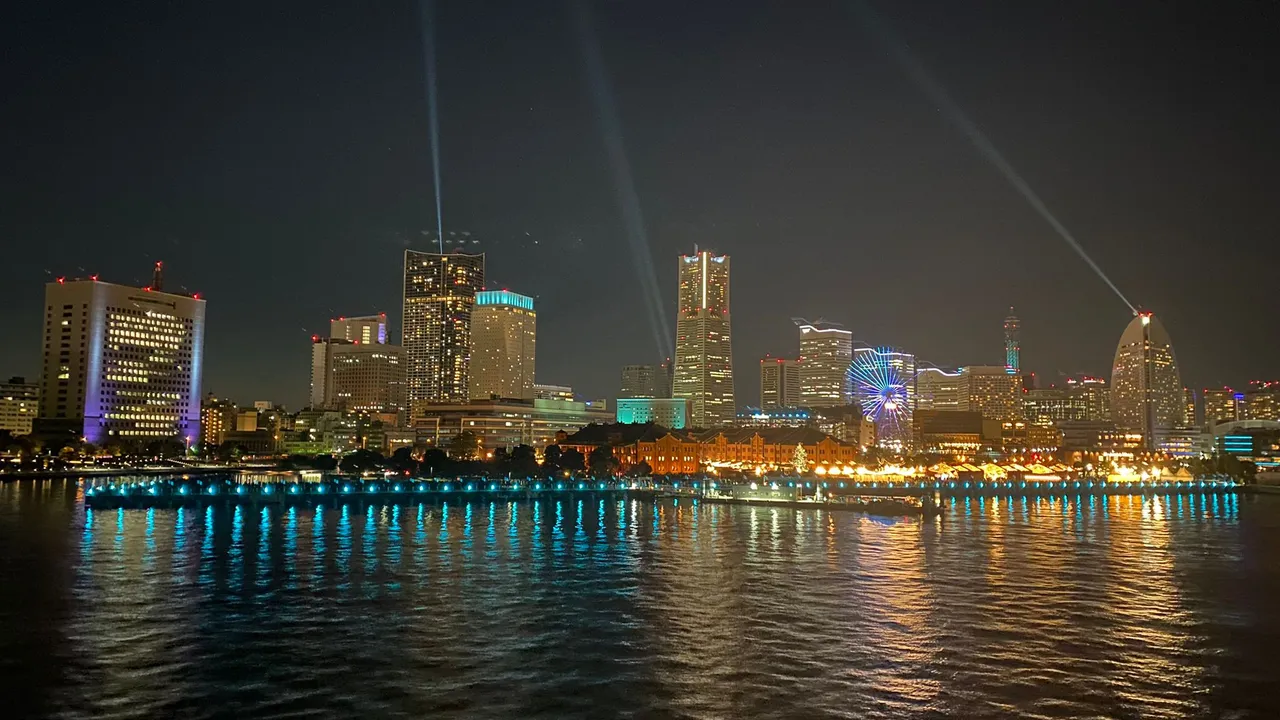 Skyline notturno di una città giapponese illuminata che si riflette sull'acqua, con fasci di luce nel cielo