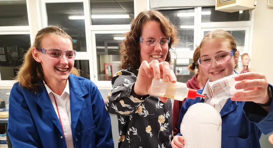 Tre studentesse del Glenfield College sono in piedi sorridenti e gesticolano in una stanza con finestre e scaffali.