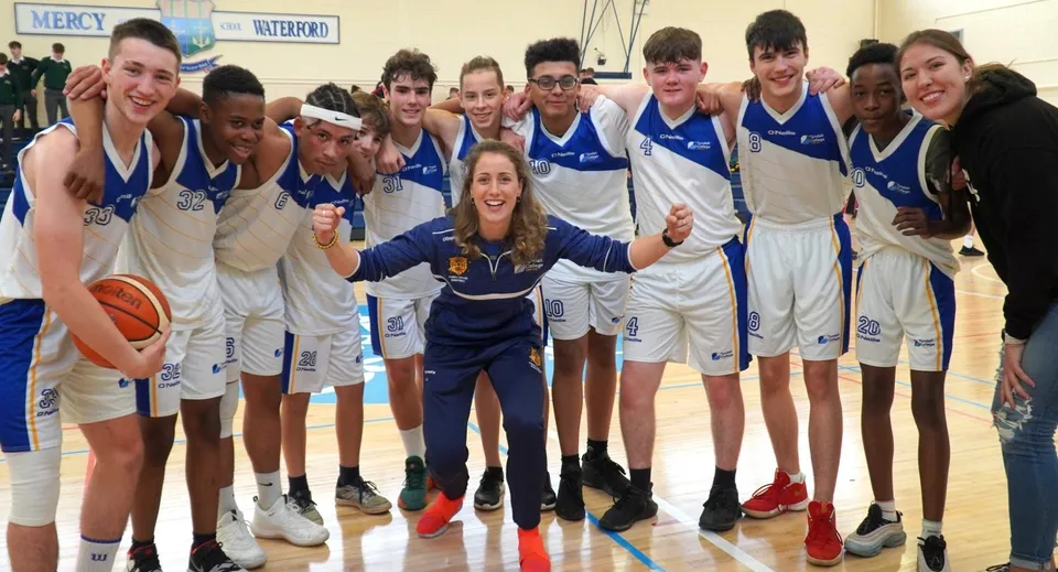 Un gruppo di giovani giocatori e giocatrici di basket del Tyndall College posa in palestra con divise blu e bianche.