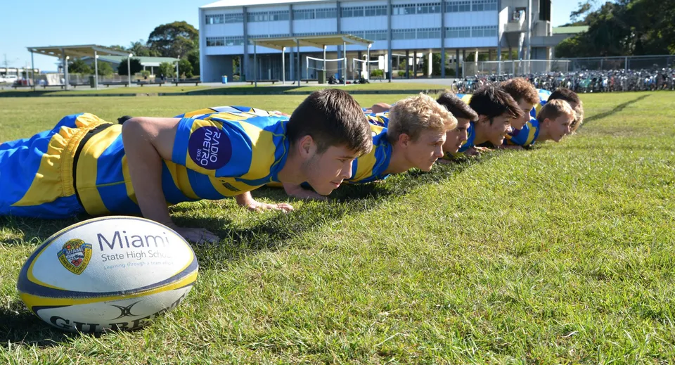 Un gruppo di studenti della Miami State High School fa flessioni su un campo sportivo davanti a una grande struttura sportiva.