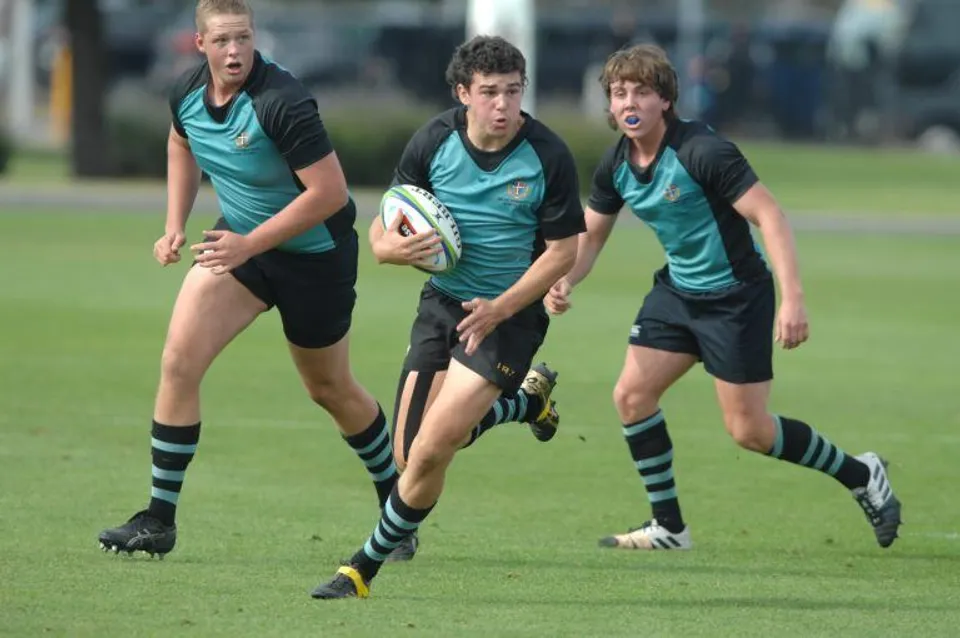 Tre giocatori di rugby della Geelong Grammar School corrono con la palla su un campo verde.