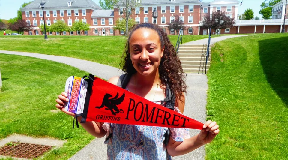 Una giovane donna sorridente regge una bandiera rosso-bianca della Pomfret sull'erba davanti a un edificio in mattoni nel campus della Pomfret School.